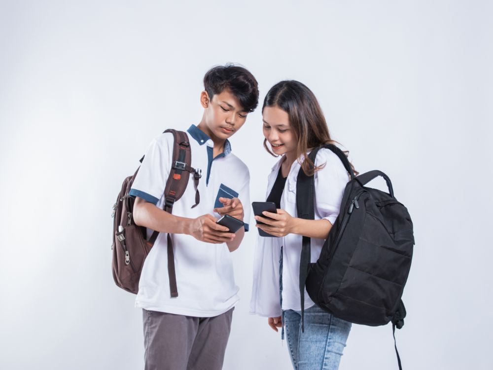 Dois estudantes com mochilas, sorrindo e compartilhando informações em seus smartphones. Imagem simbolizando a tecnologia como uma forma de captar alunos para a faculdade.