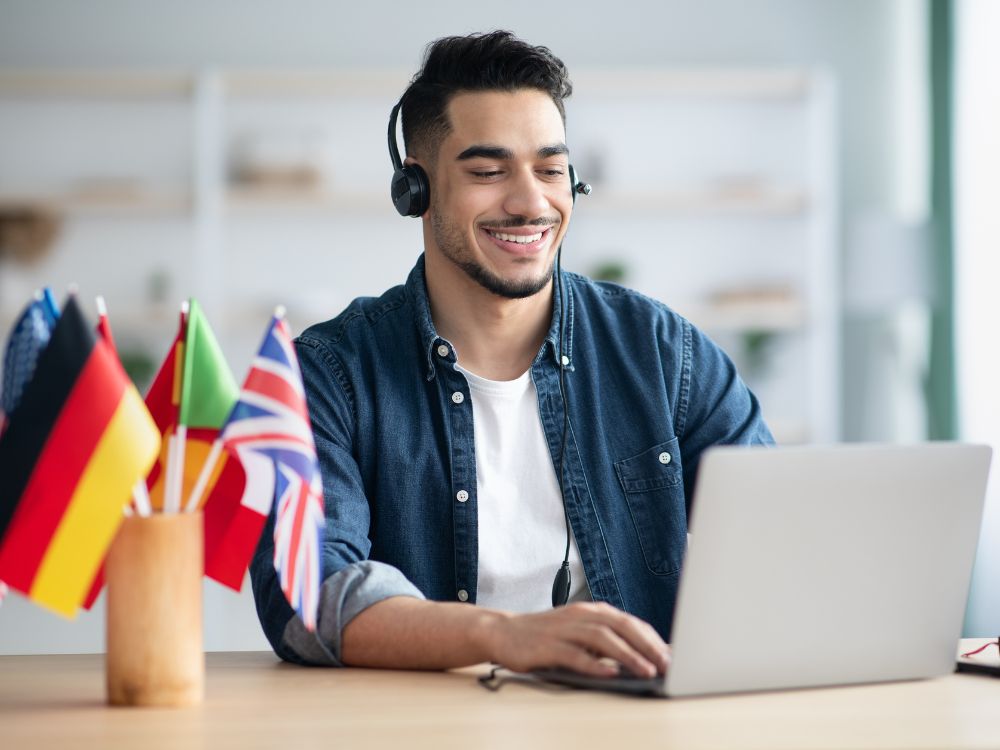 Estudante com fones de ouvido participando de aula virtual em escola de idiomas, com bandeiras representando idiomas diversos, ilustrando como captar alunos para escola de idiomas.