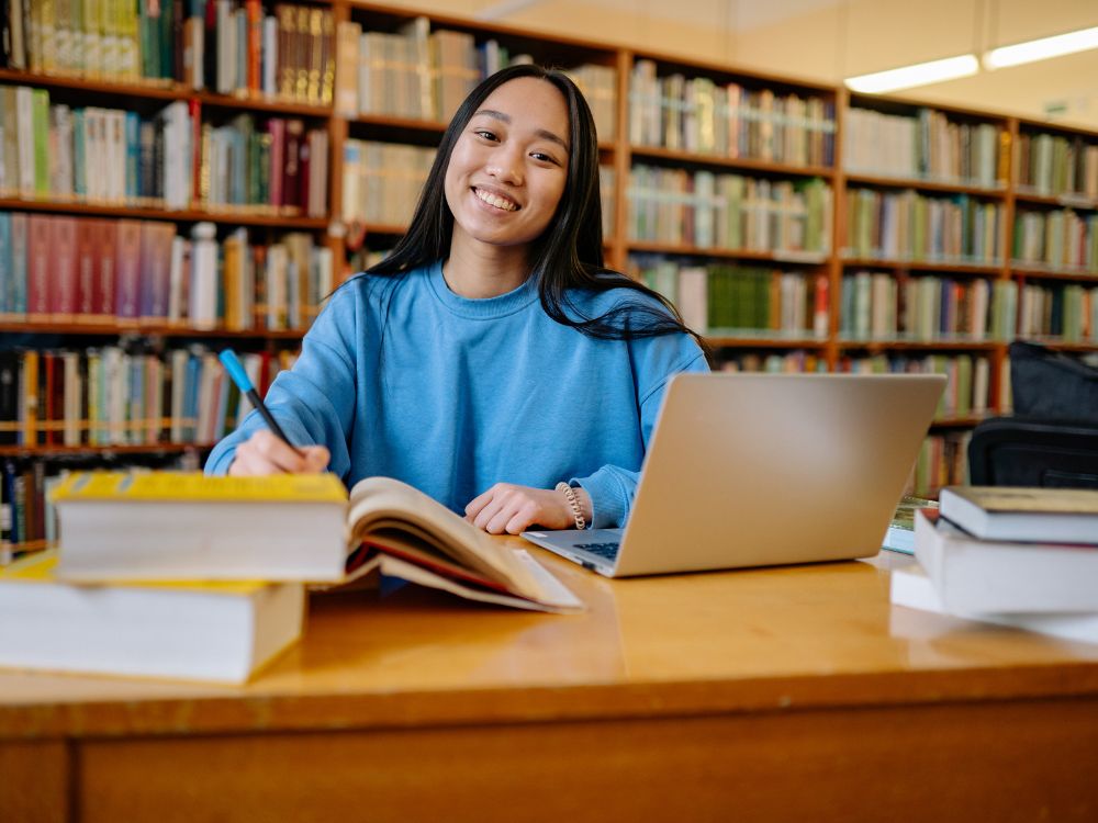 Estudante estudando em uma biblioteca com laptop e livros, destacando organização e estratégias eficazes de ações comerciais para captação de alunos.