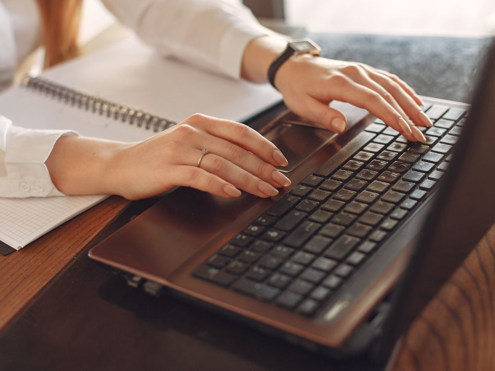 Mãos digitando em um laptop com caderno ao lado, simbolizando planejamento estratégico e ações comerciais para captação de alunos.