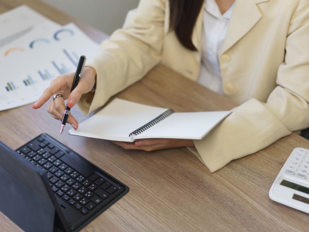 Mulher apontando para tela com caderno ao lado. Planejamento de etapas do controle de matrículas com apoio de dados e gráficos.