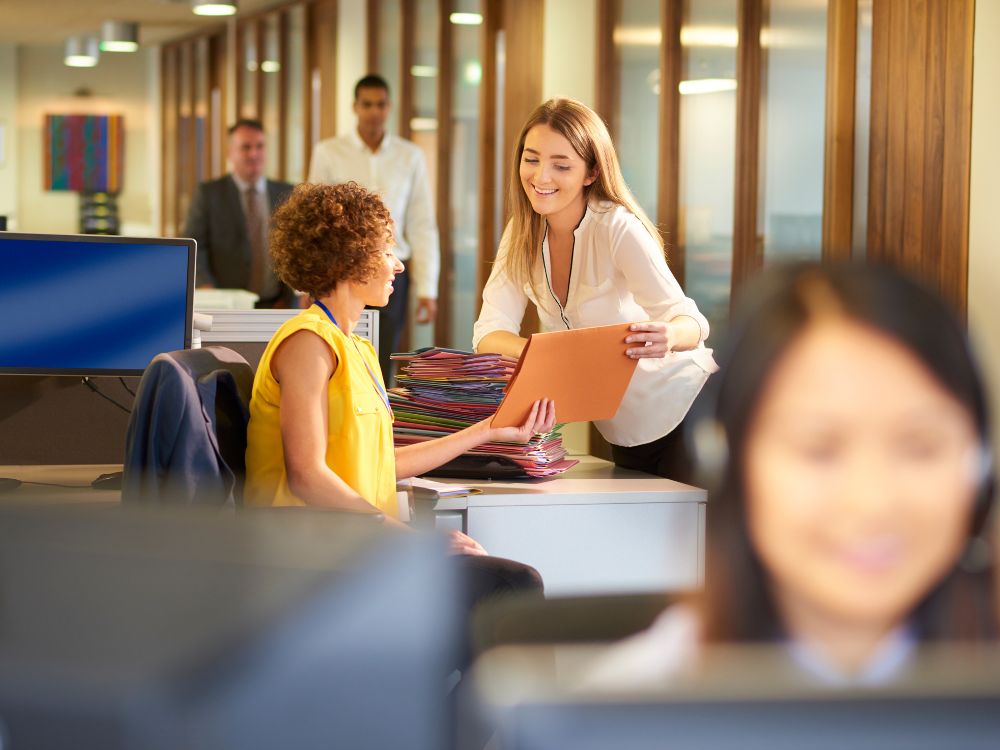 Colaboradoras trocando documentos em ambiente corporativo, simulando a organização envolvida em o que é matrícula escolar.