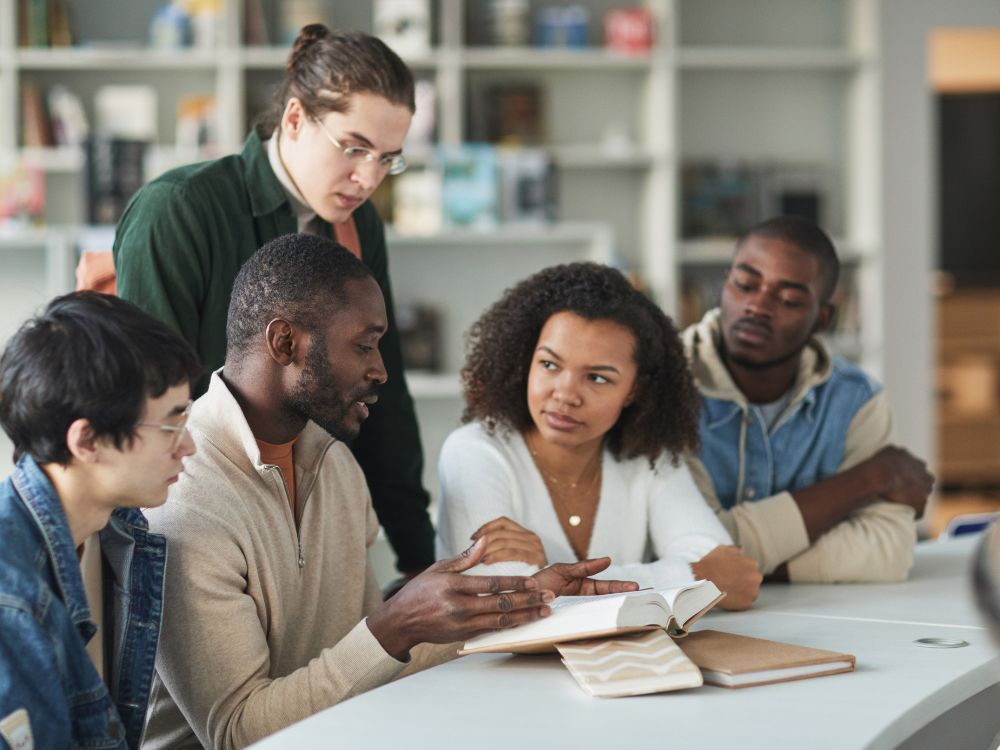 Grupo de estudantes em biblioteca discutindo livros como parte do acompanhamento acadêmico em instituições de ensino superior.