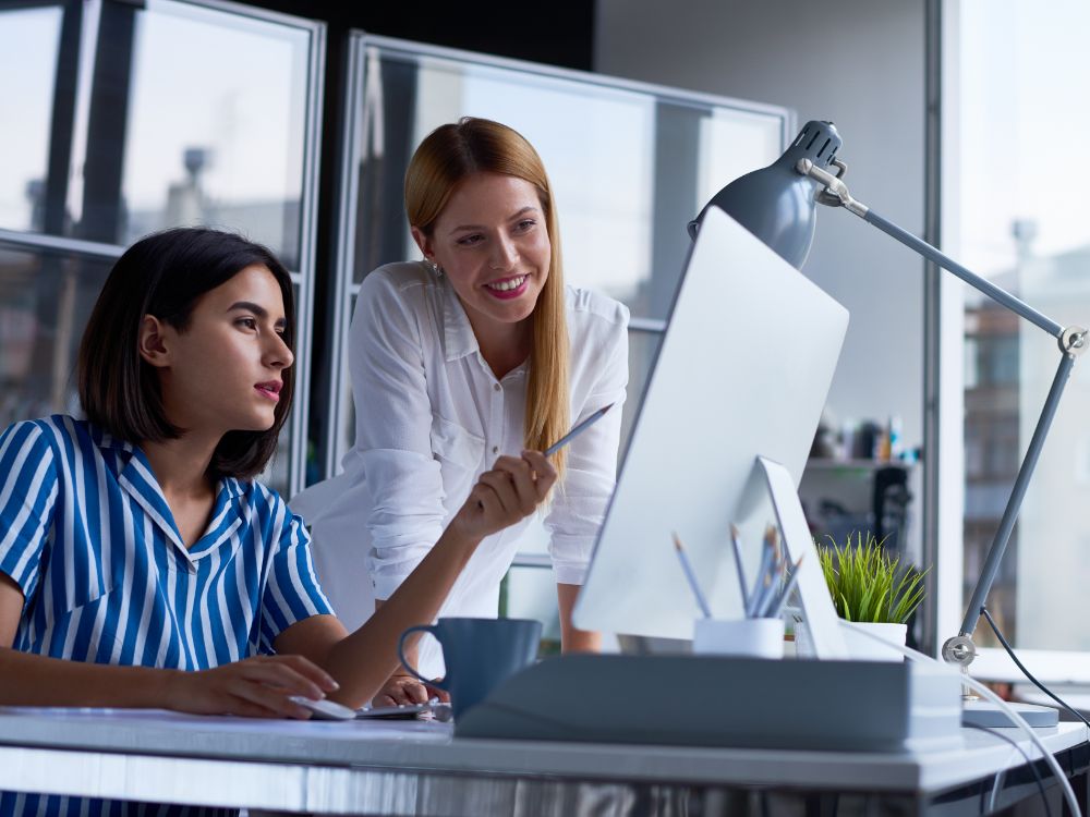 Duas profissionais observando dados na tela do computador em ambiente corporativo, representando o suporte da gestão escolar automatizada.