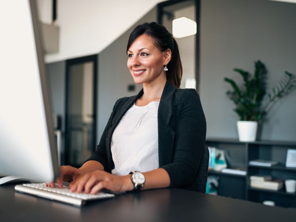Mulher digitando em computador de mesa com expressão positiva, demonstrando eficiência e organização com a gestão escolar automatizada.