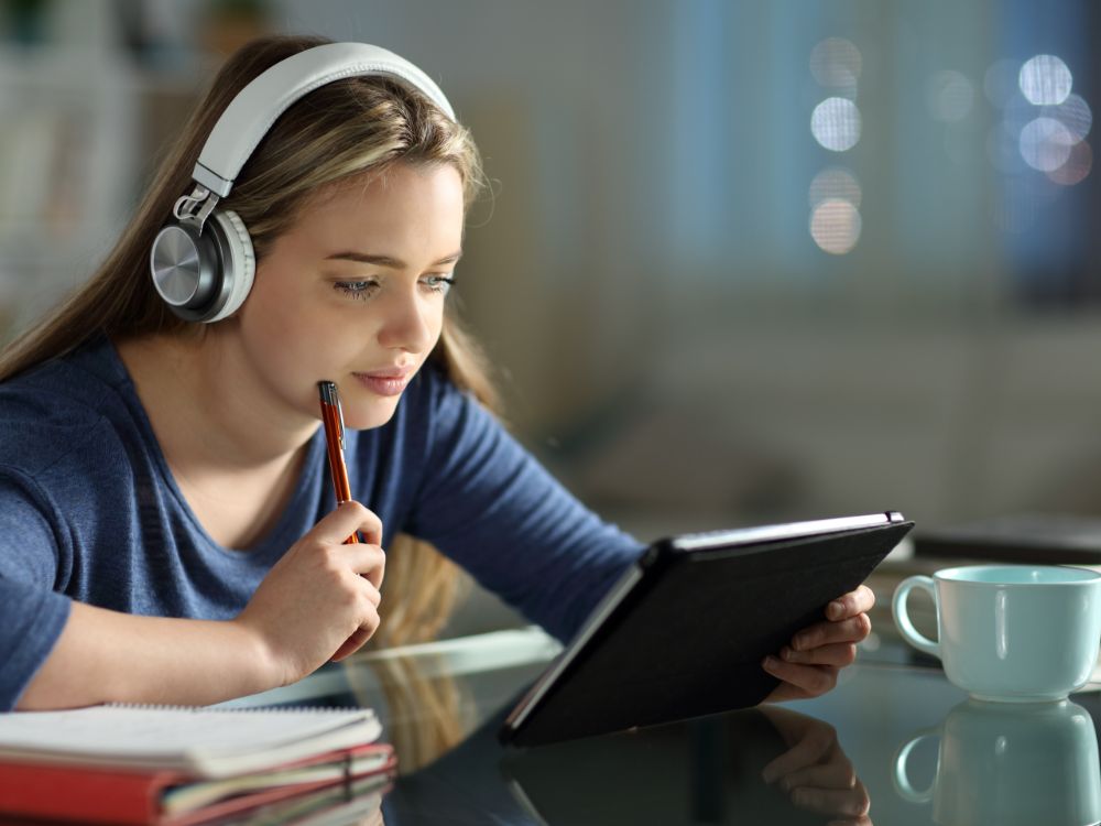 Estudante com fones de ouvido consulta material didático em tablet utilizando recursos oferecidos por uma plataforma de gestão escolar online.