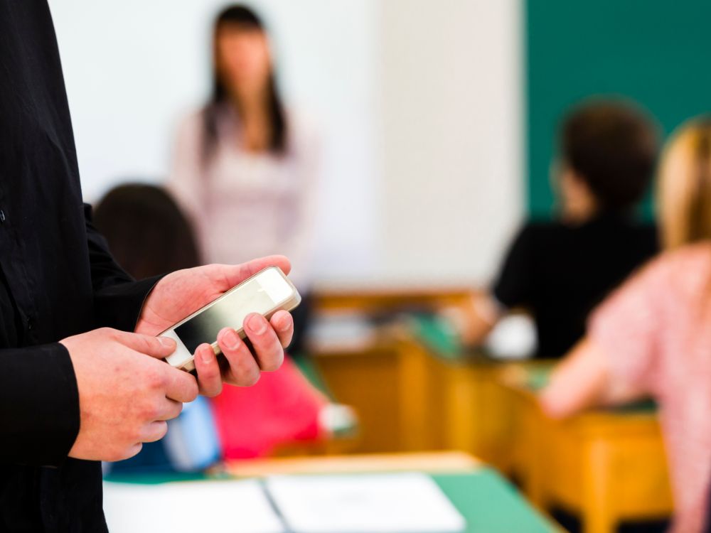 Professor segura celular em sala de aula, simbolizando o uso da Agenda Digital Escolar para registro e comunicação com os alunos.