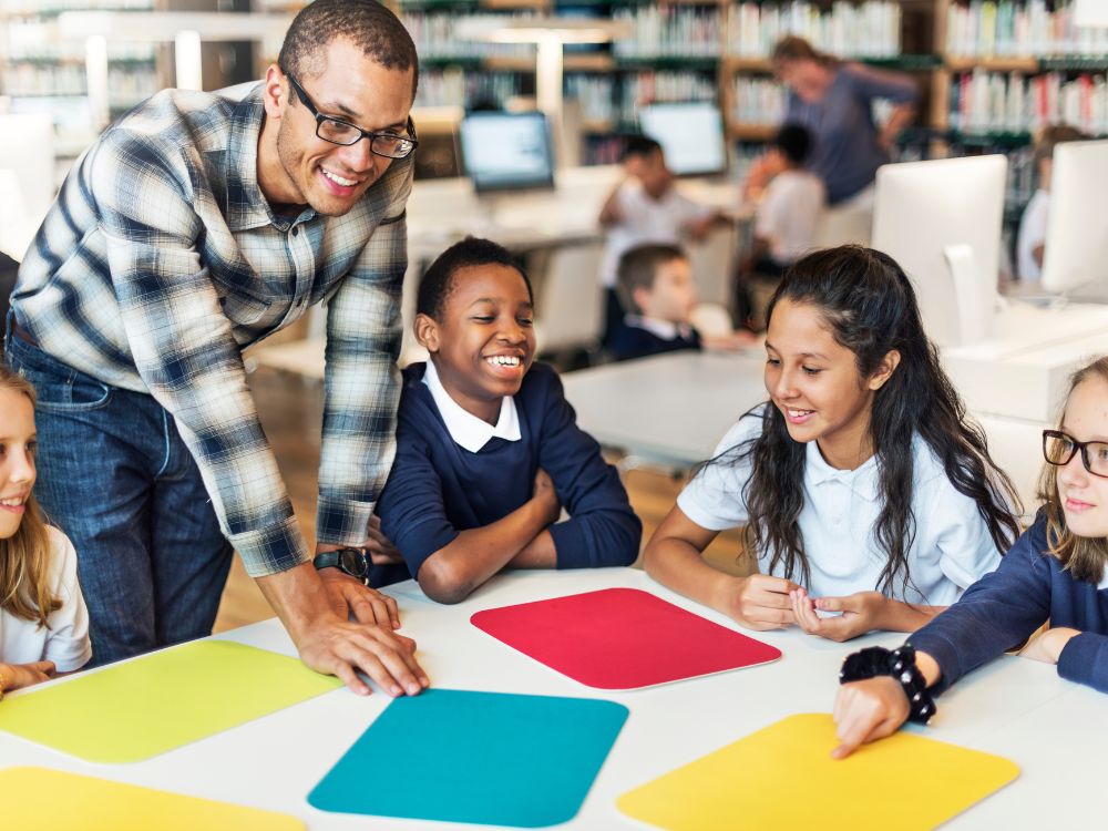 Atividade lúdica entre professor e alunos em biblioteca escolar, promovendo integração e reforçando práticas de gestão de alunos.