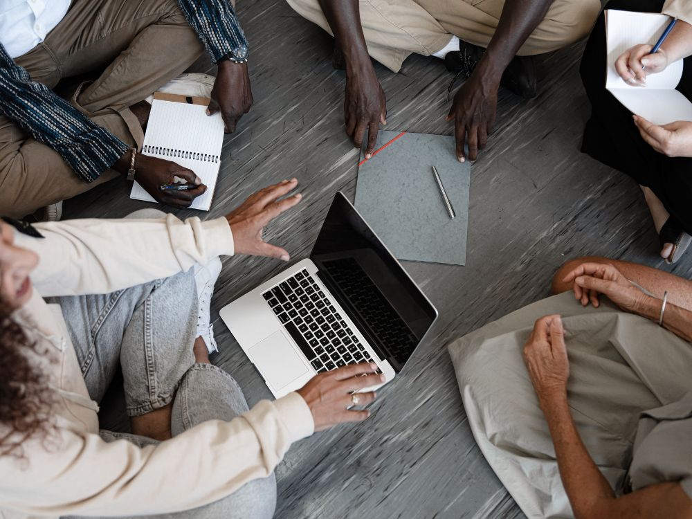 Equipe reunida no chão com anotações e notebook, discutindo formas colaborativas de organizar escola e melhorar a gestão.