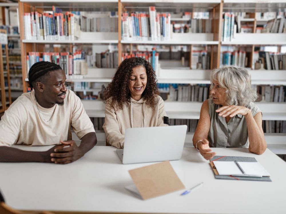 Três pessoas sorrindo em frente ao notebook em uma biblioteca, utilizando software para organizar escola de maneira prática.
