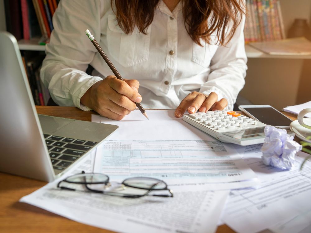 Mulher calcula e preenche planilhas financeiras com foco no plano orçamentário educacional, ao lado de notebook e documentos.