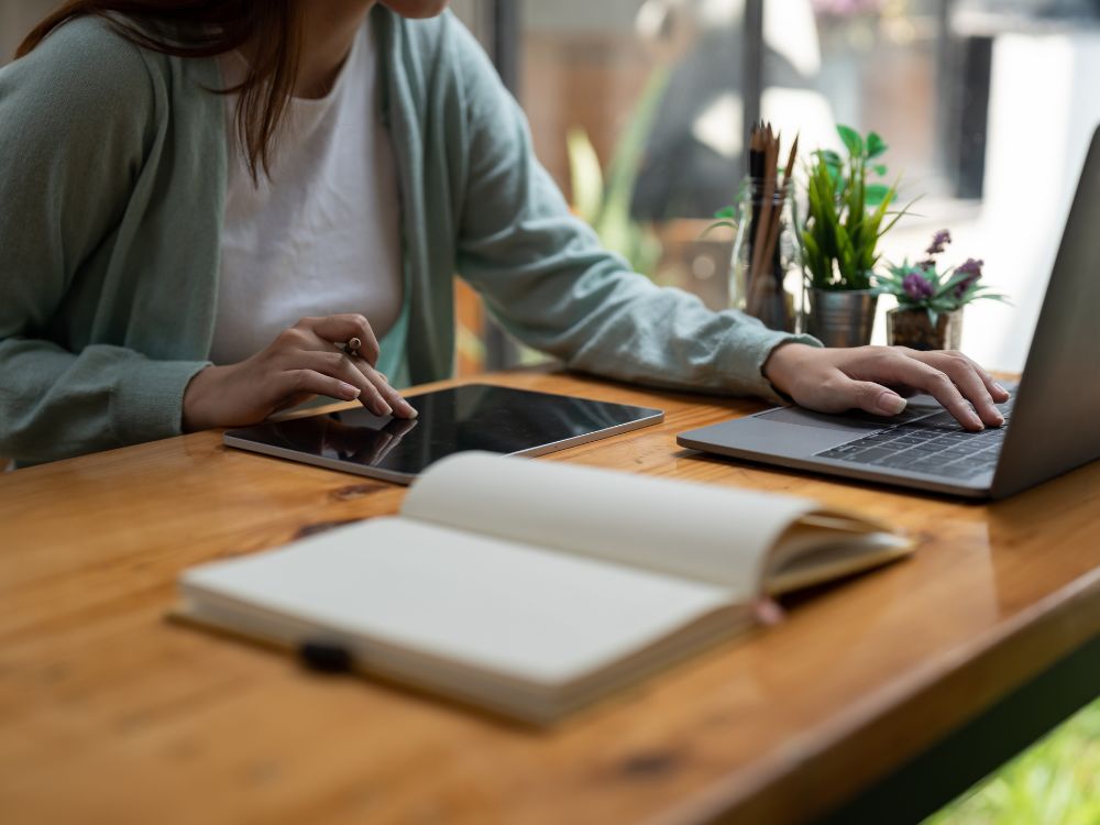 Estudante utilizando notebook e tablet sobre mesa de estudos, com caderno aberto e materiais de escritório, representando o uso de software para faculdade no apoio às atividades acadêmicas.