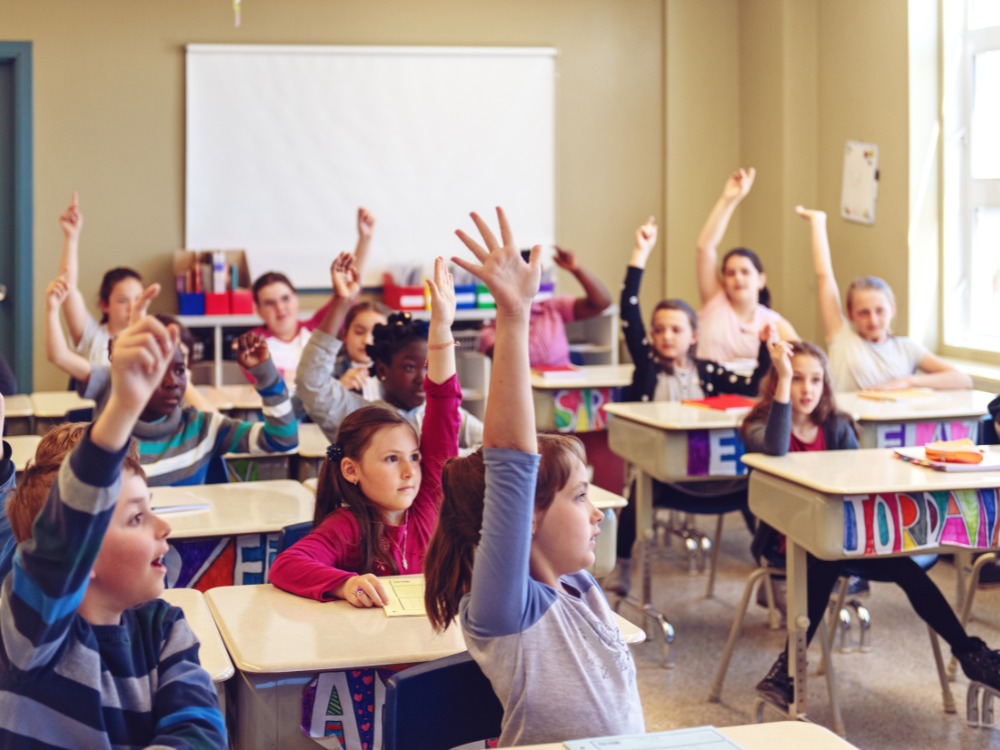 Alunos levantam as mãos em uma sala de aula para responder a uma pergunta do professor. Esta imagem ilustra a participação e o engajamento que podem ser alcançados com um bom gerenciamento de alunos.