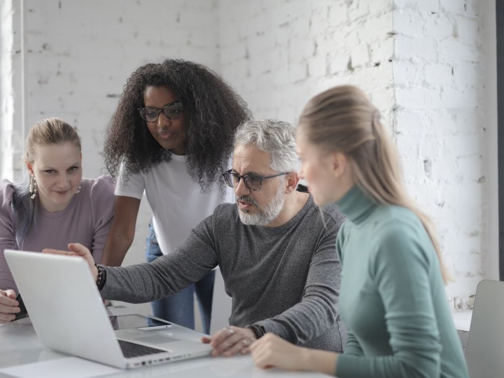 Quatro profissionais, incluindo um homem de cabelo grisalho, em uma reunião de equipe em volta de um notebook. Eles discutem um projeto em um escritório.