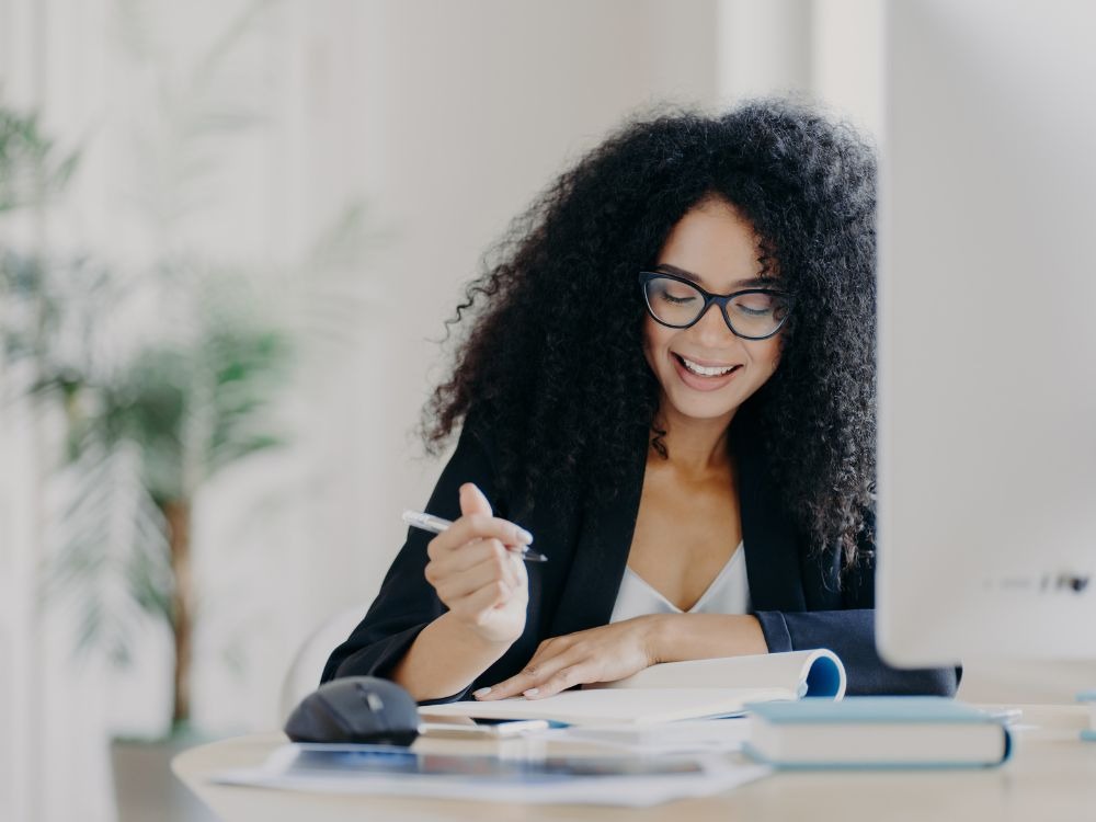 Mulher negra com cabelo cacheado e óculos, sorrindo e anotando algo em um caderno enquanto está em sua mesa de trabalho com um computador.
