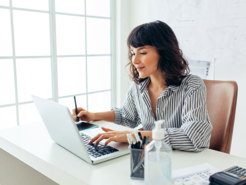 Professora utilizando software para faculdade em um laptop, em ambiente de estudo com materiais organizados e tela clara ao fundo.