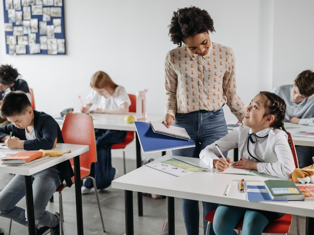 Professora de blusa florida e calça jeans auxilia uma aluna em uma carteira de escola. O cenário mostra uma sala de aula e representa a atenção individual que um bom gerenciamento de alunos pode oferecer.