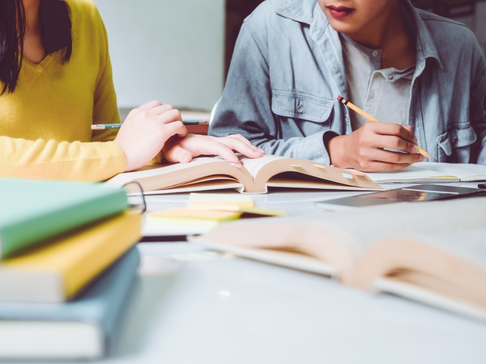 Duas pessoas sentadas em uma mesa de estudo, lendo livros e anotando, mostrando dedicação ao aprendizado. A cena simboliza o foco e a produtividade no estudo, resultado de um bom gerenciamento de alunos.