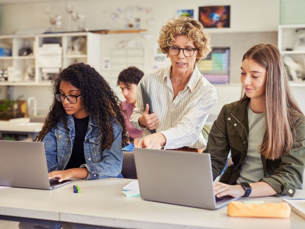Professora auxilia alunas em atividades com notebook, mostrando como a tecnologia contribui para o acompanhamento dos indicadores educacionais.