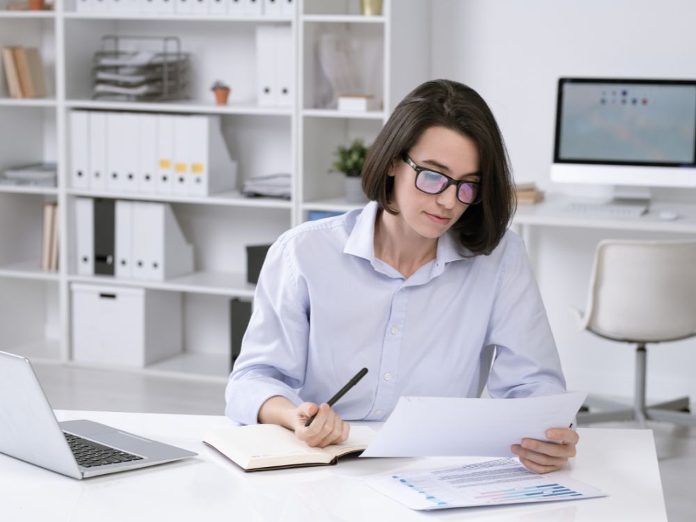 Mulher lendo relatórios e fazendo anotações em um ambiente de trabalho, simbolizando a importância do planejamento e da organização escolar.