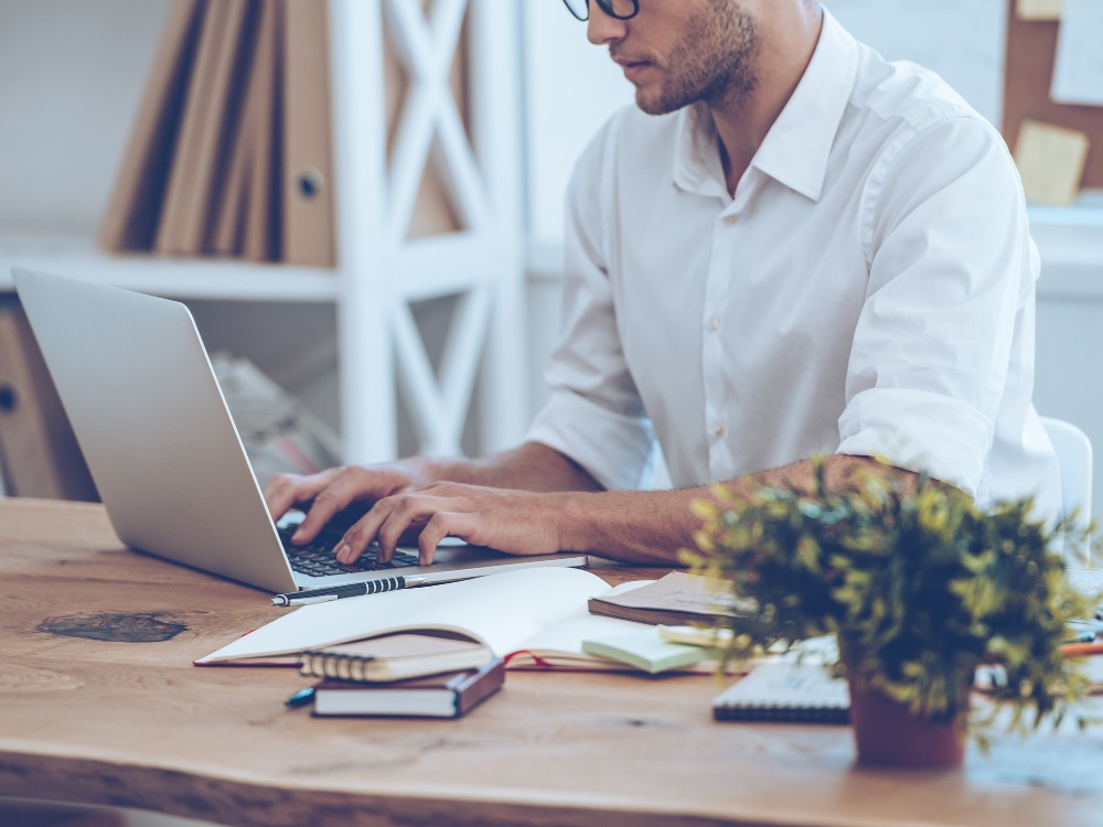 Homem trabalhando em um notebook em um escritório moderno, digitando em um ambiente organizado com cadernos e planta sobre a mesa e fazendo analise de dados educacionais.
