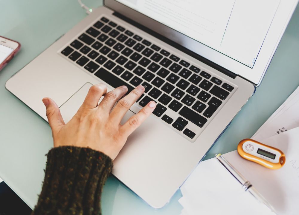 Mão de pessoa digitando em teclado de notebook em ambiente de trabalho com documentos ao lado – conceito de trabalho digital, produtividade e uso da tecnologia.