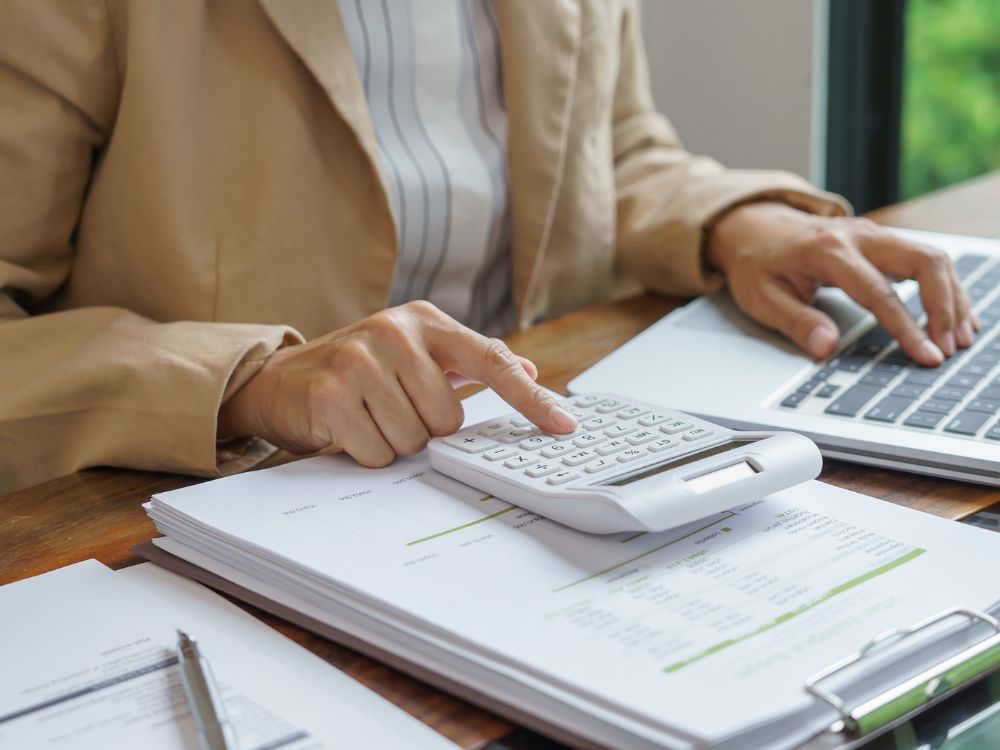 Pessoa calculando despesas e receitas com planilhas e calculadora, destacando o controle essencial na gestão financeira escolar.