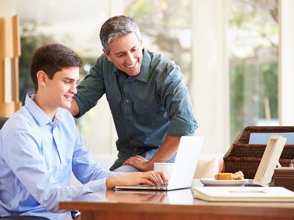 Pai auxiliando o filho em atividade online no notebook, simbolizando o acesso ao portal do aluno e responsável para acompanhamento escolar.