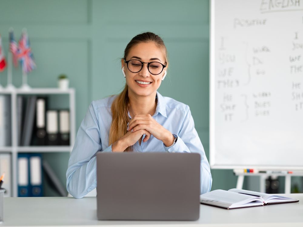 Mulher sorridente participando de reunião online, com laptop aberto e quadro com anotações de aula ao fundo.