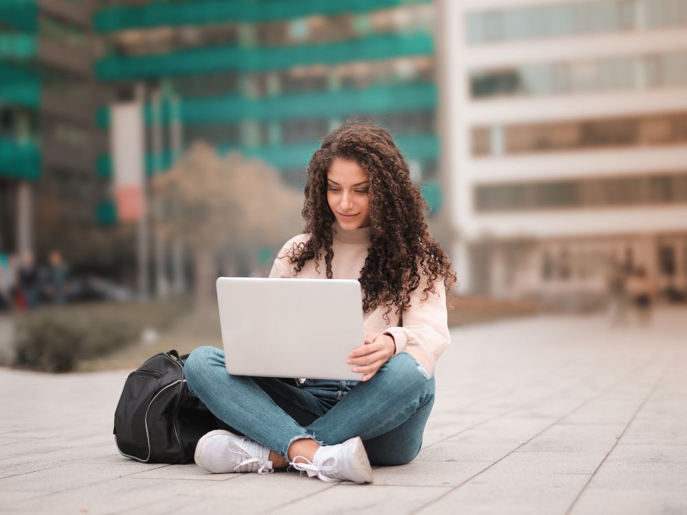 Jovem sentada no chão em área externa, usando notebook no colo, com mochila ao lado e prédios ao fundo.