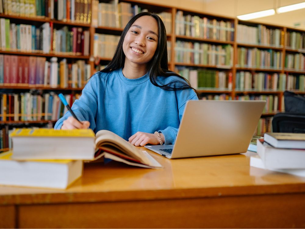 Estudante sorrindo enquanto faz anotações em um livro aberto, sentada em mesa de biblioteca com notebook e pilhas de livros ao redor.
