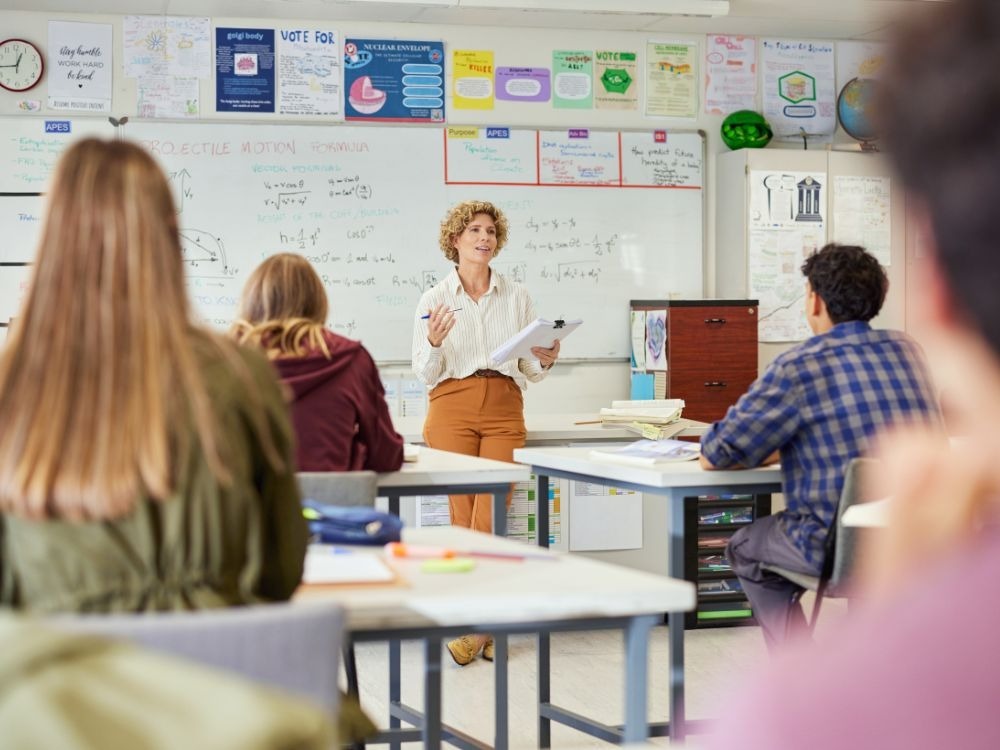 Professora conduzindo aula em sala, demonstrando como avaliar os alunos com métodos pedagógicos e acompanhamento em sala de aula.