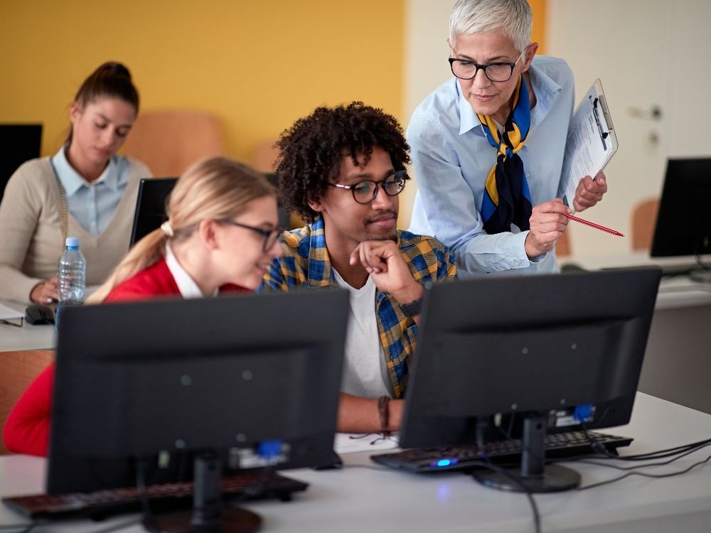 Equipe educacional orientando alunos em laboratório de informática, ilustrando como avaliar os alunos por meio de atividades práticas e colaborativas.