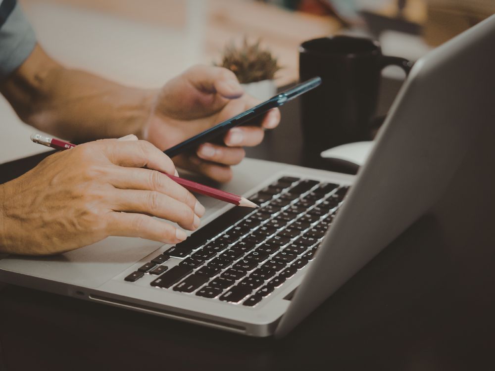 Profissional utilizando notebook e celular para atualização de dados acadêmicos, representando o Ajuste de Matrículas em sistemas educacionais.