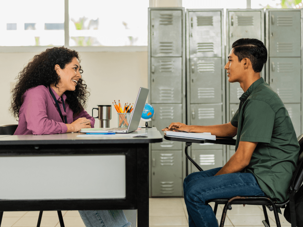 Funcionária da escola conversa com aluno em mesa de atendimento, representando o suporte da secretaria escolar.