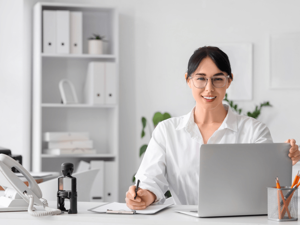Funcionária trabalha em notebook com bloco de anotações sobre a mesa, simbolizando a administração escolar.