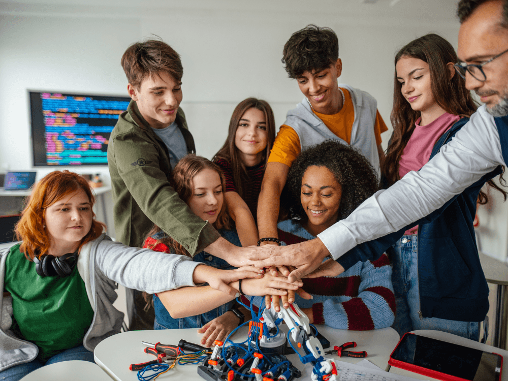 Estudantes e professor unem as mãos durante atividade colaborativa com robótica, representando o que é inclusão escolar no ambiente educacional.
